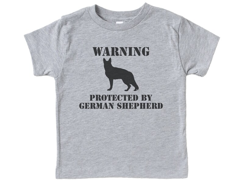 Pu&ograve; includere: Maglietta grigia a maniche corte con una silhouette nera di un pastore tedesco e il testo "WARNING PROTECTED BY GERMAN SHEPHERD".