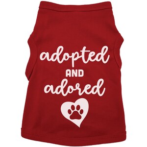 May include: Red sleeveless dog shirt with white text that says "adopted and adored" with a white paw print inside a heart.
