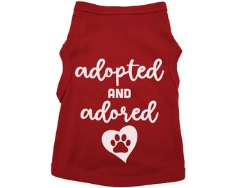 May include: Red sleeveless dog shirt with white text that says "adopted and adored" with a white paw print inside a heart.