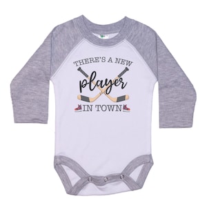 May include: White long-sleeve baby bodysuit with a grey raglan sleeve design. The bodysuit has a graphic of two hockey sticks crossed with the text "There's a new player in town".