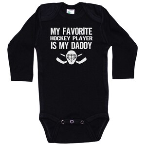 May include: Black long-sleeve baby onesie with white text that reads "My favorite hockey player is my daddy" and a hockey stick and goalie mask graphic.