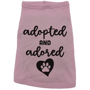 May include: Pink dog shirt with black text that says "adopted and adored" with a black paw print inside a heart.