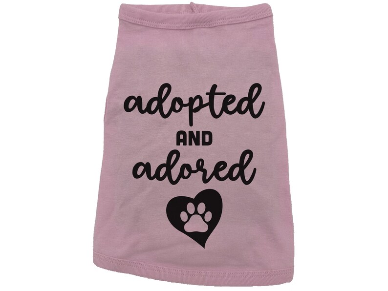 May include: Pink dog shirt with black text that says "adopted and adored" with a black paw print inside a heart.