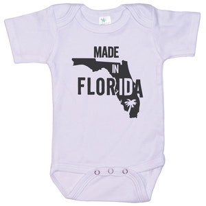 K&ouml;nnte beinhalten: Ein wei&szlig;er Baby-Strampler mit einer schwarzen Umrandung des Bundesstaates Florida und dem Text "Made in Florida".