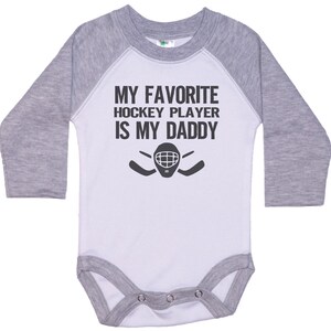 May include: A white and gray long-sleeve baby bodysuit with a black graphic that says "My favorite hockey player is my daddy" with a hockey mask and sticks.