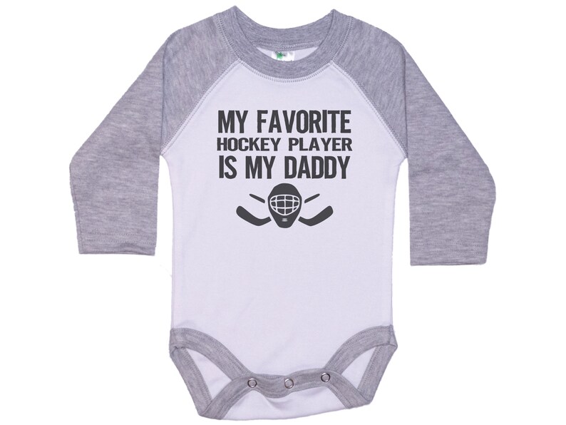 May include: A white and gray long-sleeve baby bodysuit with a black graphic that says "My favorite hockey player is my daddy" with a hockey mask and sticks.