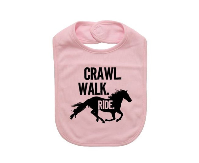 May include: A light pink baby bib with a black graphic of a running horse and the words "Crawl. Walk. Ride." printed in black. The bib has a snap closure at the neck.