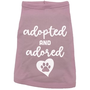 May include: Pink dog shirt with white text that says "adopted and adored" with a white paw print inside a heart.