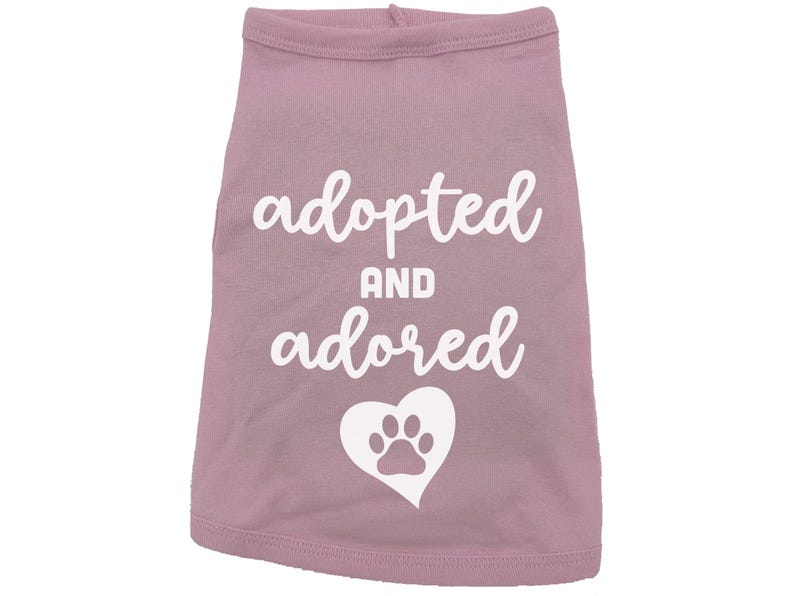 May include: Pink dog shirt with white text that says "adopted and adored" with a white paw print inside a heart.