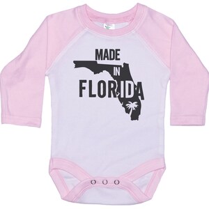 K&ouml;nnte beinhalten: Wei&szlig;er Langarm-Babybody mit rosa &Auml;rmeln. Der Body hat einen schwarzen Umriss des Bundesstaates Florida mit dem aufgedruckten Text "MADE IN FLORIDA".