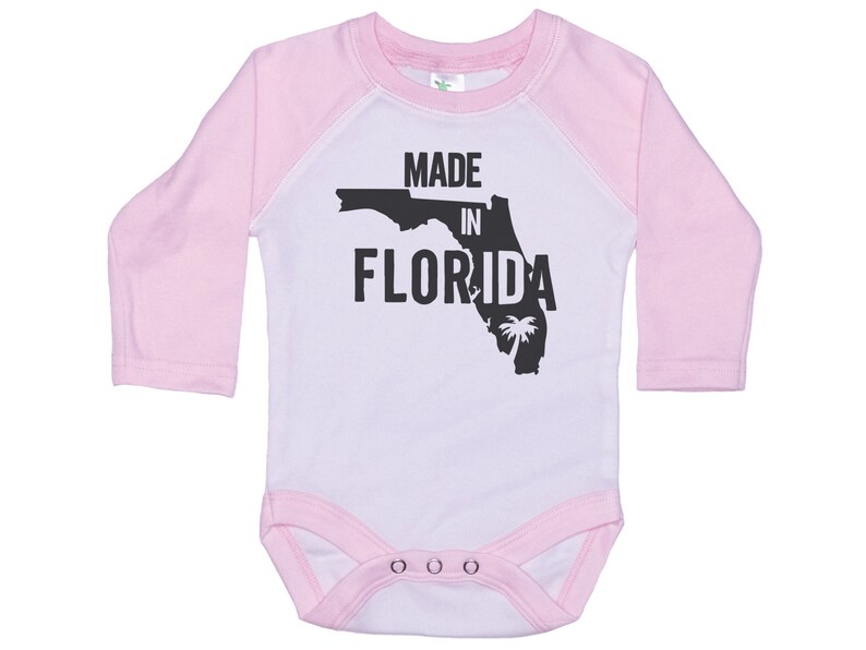 K&ouml;nnte beinhalten: Wei&szlig;er Langarm-Babybody mit rosa &Auml;rmeln. Der Body hat einen schwarzen Umriss des Bundesstaates Florida mit dem aufgedruckten Text "MADE IN FLORIDA".
