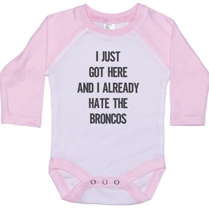May include: Pink and white long-sleeved baby onesie with the text "I just got here and I already hate the Broncos".