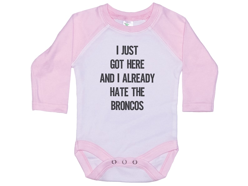 May include: Pink and white long-sleeved baby onesie with the text "I just got here and I already hate the Broncos".