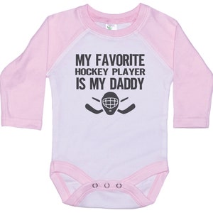May include: White long-sleeve baby onesie with pink sleeves. The onesie has a black graphic that says "My favorite hockey player is my daddy" with a hockey stick and goalie mask.