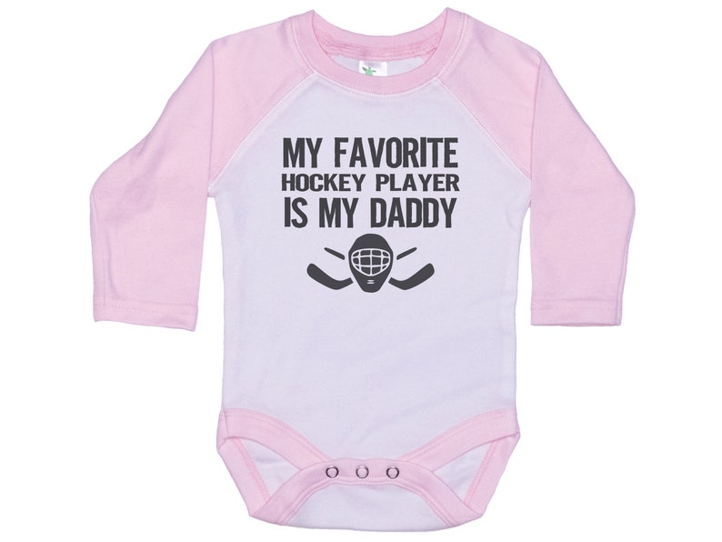 May include: White long-sleeve baby onesie with pink sleeves. The onesie has a black graphic that says "My favorite hockey player is my daddy" with a hockey stick and goalie mask.
