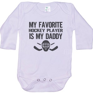 May include: White long-sleeved baby onesie with black text that reads "My favorite hockey player is my daddy" and a hockey mask and stick graphic.