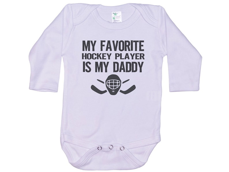 May include: White long-sleeved baby onesie with black text that reads "My favorite hockey player is my daddy" and a hockey mask and stick graphic.