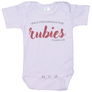 May include: A white baby onesie with short sleeves and a pink glitter design that says "she is more precious than rubies Proverbs 3:15".