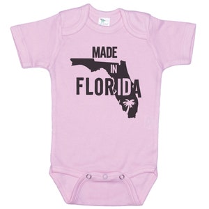 K&ouml;nnte beinhalten: Rosaer Baby-Strampler mit einer schwarzen Umrandung des Bundesstaates Florida und dem Text "Made in Florida".