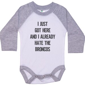 May include: A white and gray long-sleeve baby onesie with the text "I just got here and I already hate the Broncos" printed on the front.