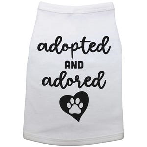 May include: White dog shirt with black text that says "adopted and adored" with a black heart and white paw print inside.