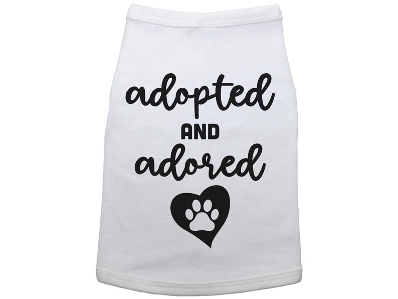 May include: White dog shirt with black text that says "adopted and adored" with a black heart and white paw print inside.