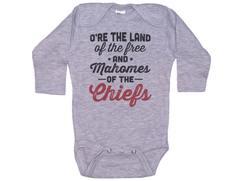 Pode incluir: Macac&atilde;o de beb&ecirc; de manga comprida cinza com o texto "O're the land of the free and Mahomes -of the- Chiefs" em preto e vermelho.