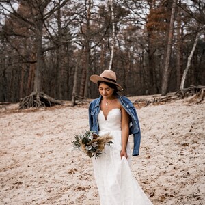 Pode incluir: Uma noiva usando um vestido de noiva de renda branca e uma jaqueta de ganga caminha por uma floresta. Ela est&aacute; usando um chap&eacute;u marrom e carregando um buqu&ecirc; de flores.