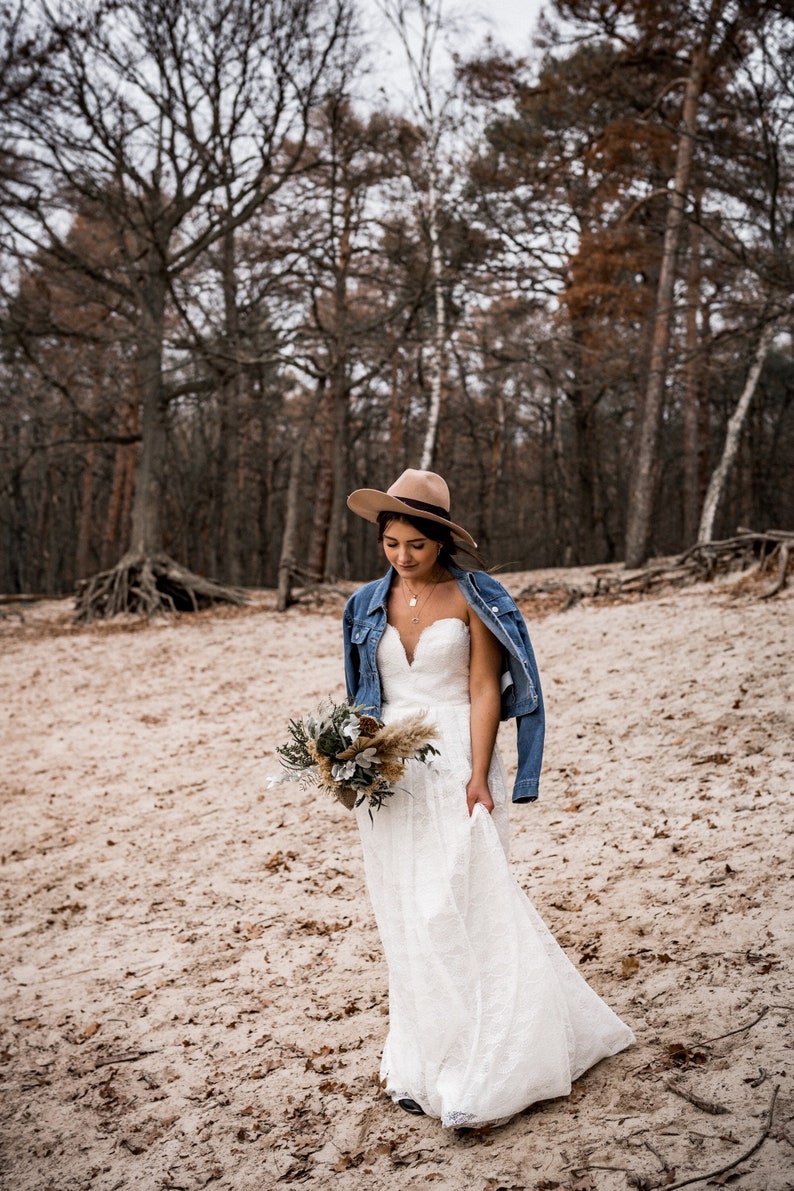 Pode incluir: Uma noiva usando um vestido de noiva de renda branca e uma jaqueta de ganga caminha por uma floresta. Ela est&aacute; usando um chap&eacute;u marrom e carregando um buqu&ecirc; de flores.