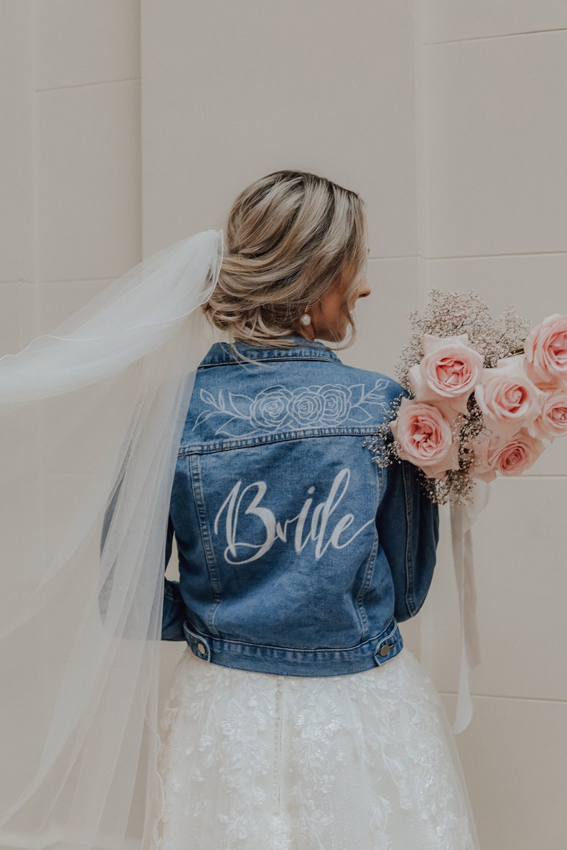 Pode incluir: Uma noiva usando um v&eacute;u branco e uma jaqueta de ganga com a palavra "Bride" escrita nas costas em letras brancas. Ela est&aacute; segurando um buqu&ecirc; de rosas cor-de-rosa.