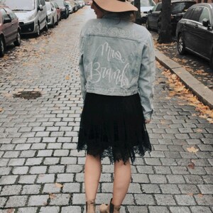 Pode incluir: Uma mulher vestindo uma jaqueta de jeans azul claro com as palavras "Mrs. Brand" escritas em branco nas costas. Ela tamb&eacute;m est&aacute; vestindo uma saia preta, um chap&eacute;u marrom e botas de cano baixo marrons. Ela est&aacute; caminhando por uma rua de paralelep&iacute;pedos.