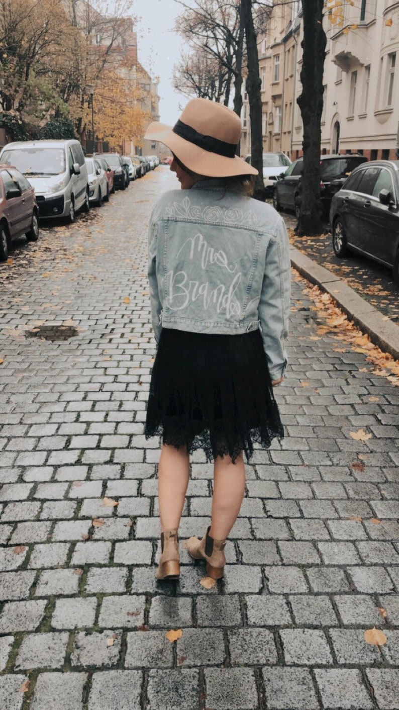 Pode incluir: Uma mulher vestindo uma jaqueta de jeans azul claro com as palavras "Mrs. Brand" escritas em branco nas costas. Ela tamb&eacute;m est&aacute; vestindo uma saia preta, um chap&eacute;u marrom e botas de cano baixo marrons. Ela est&aacute; caminhando por uma rua de paralelep&iacute;pedos.