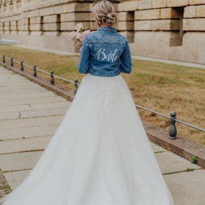 Pode incluir: Uma noiva usando um vestido de noiva branco e uma jaqueta jeans com a palavra "Bride" escrita nas costas. Ela est&aacute; segurando um buqu&ecirc; de flores rosa.
