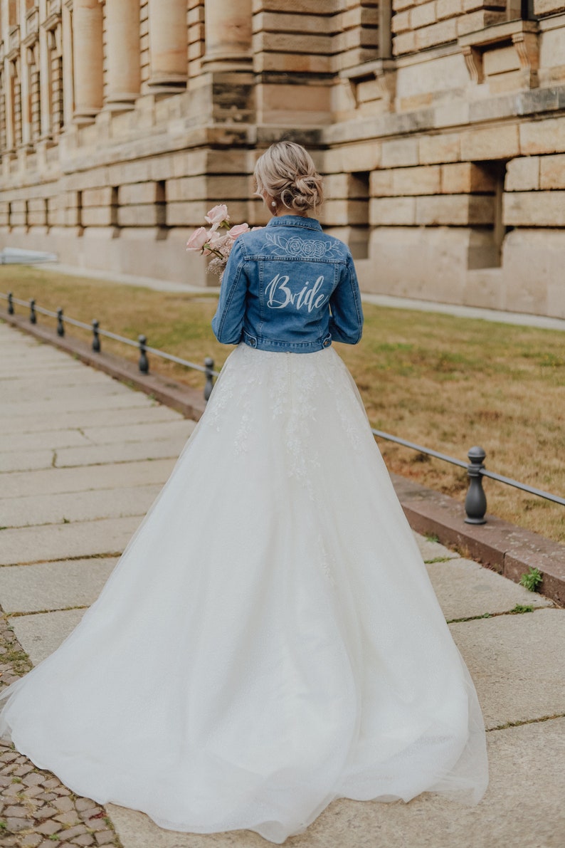 Pode incluir: Uma noiva usando um vestido de noiva branco e uma jaqueta jeans com a palavra "Bride" escrita nas costas. Ela est&aacute; segurando um buqu&ecirc; de flores rosa.