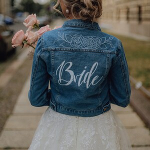 Pode incluir: Uma noiva usando um vestido de noiva branco com uma jaqueta de ganga que diz "Bride" nas costas. A jaqueta tem um design floral com tr&ecirc;s rosas nas costas.