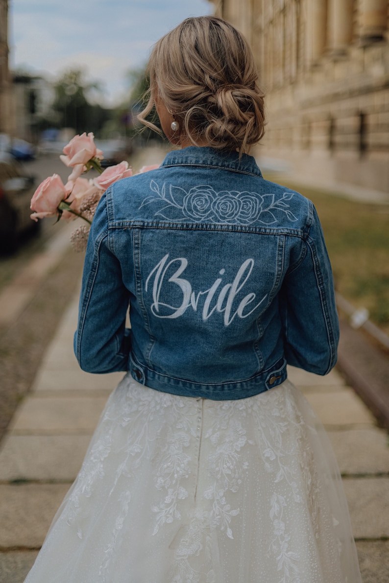 Pode incluir: Uma noiva usando um vestido de noiva branco com uma jaqueta de ganga que diz "Bride" nas costas. A jaqueta tem um design floral com tr&ecirc;s rosas nas costas.