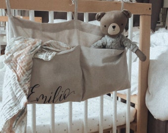 May include: A beige fabric crib organizer with two pockets, one with the name "Emilio" printed on it. A brown teddy bear is in the top pocket. The organizer is hanging on a white crib with a white and gold polka dot sheet.