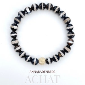 May include: Black and white agate gemstone bracelet with a gold bead accent. ANNABADENBERG ACHAT.