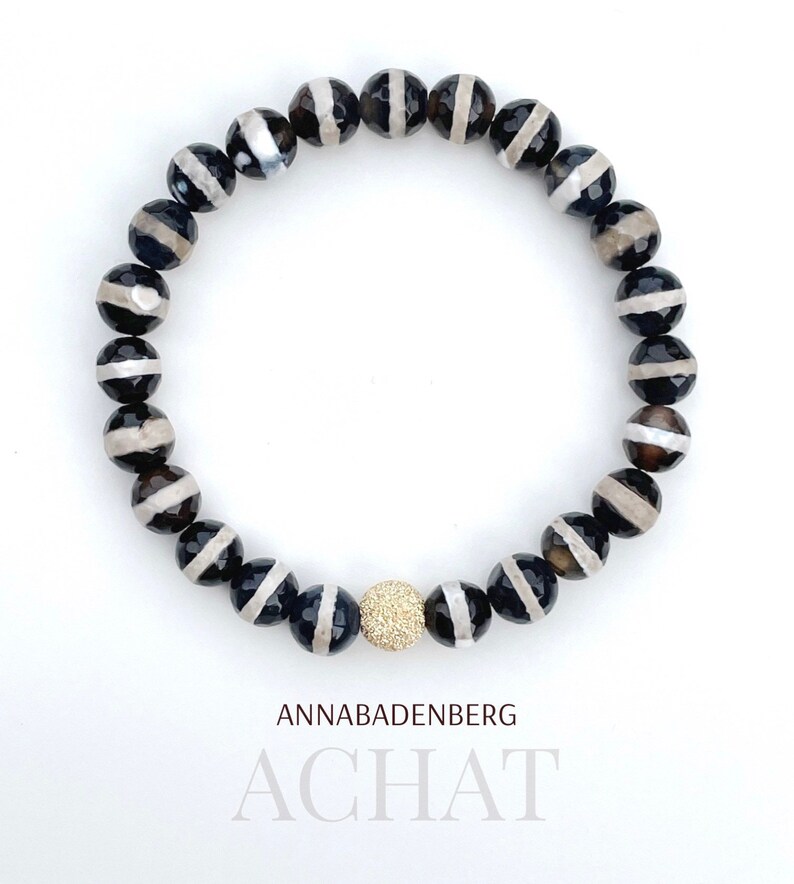 May include: Black and white agate gemstone bracelet with a gold bead accent. ANNABADENBERG ACHAT.