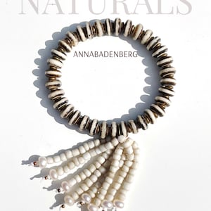 Könnte beinhalten: Ein weißes und braunes Perlenarmband mit einem weißen Quastenanhänger. Das Armband trägt den Text "ANNABADENBURG". Das Armband liegt auf einem weißen Hintergrund.
