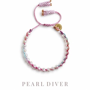 May include: A bracelet featuring white pearls strung on pink and light blue cord, with two light pink beaded tassels at the ends. A small gold-toned circular charm is attached. The text PEARL DIVER MADE BY ANNA BADENBERG is printed below.