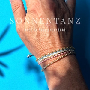 May include: Two bracelets featuring gold-tone beads and a woven pattern in light blue, white, and pink. The text SONNENTANZ MADE BY ANNA BADENBERG is visible. The bracelets are adjustable with a sliding knot closure and small dangling beads.