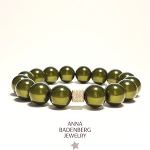 Könnte beinhalten: Ein grünes Perlenarmband mit einem goldenen quadratischen Anhänger, der mit klaren Steinen besetzt ist. Das Armband besteht aus runden, glänzenden Perlen.