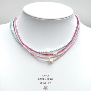 Könnte beinhalten: Eine mehrreihige Perlenkette mit hellblauen, dunkelrosa, hellrosa und klaren Glasperlen. Die Kette ist mit drei weißen Perlen unterschiedlicher Größe verziert. Unter der Kette steht der Text ANNA BADENBERG JEWELRY mit einem Stern.