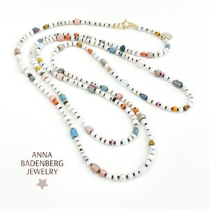 Può includere: Una collana multifilo con perline bianche e accenti colorati. La collana presenta un mix di perline rotonde e rettangolari in tonalità di blu, rosa, arancio e giallo. Una singola perla è visibile al centro della collana.