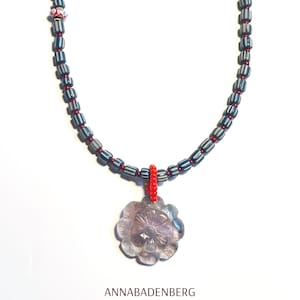 Puede incluir: Un collar hecho de cuentas rayadas azules y blancas con un acento de cuentas rojas. El collar tiene un colgante de flor de amatista tallada.