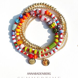 Puede incluir: Un conjunto de pulseras de cuentas coloridas con detalles dorados. Las pulseras presentan una variedad de cuentas de vidrio en rojo, naranja, amarillo, verde, azul y blanco. Las pulseras están apiladas una encima de la otra y tienen pequeños dijes dorados con el texto "Anna Badenberg" en ellos.