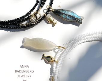 May include: Two beaded necklaces with fish pendants. One necklace is black with a labradorite fish pendant and a gold fish charm. The other is white with a mother-of-pearl fish pendant and a gold fish charm. The necklaces have gold clasps.