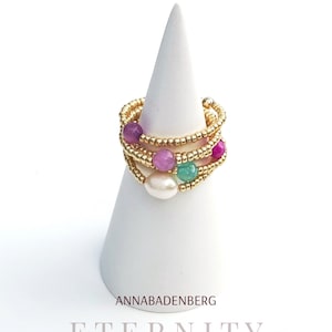 Könnte beinhalten: Goldring mit mehreren Bändern aus kleinen goldenen Perlen und bunten Steinen. Die Steine sind rosa, lila, grün und weiß. Der Ring befindet sich auf einem weißen, kegelförmigen Ringständer. Der Text "ANNABADENBERG" und "ETERNITY" befindet sich unter dem Ring.