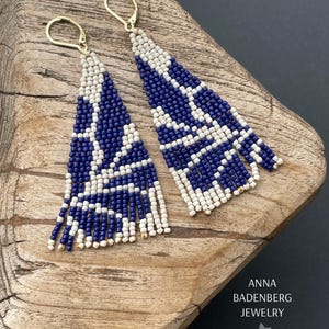 Könnte beinhalten: Ein Paar lange, geperlte Ohrringe mit einem geometrischen Muster in Weiß und Marineblau. Die Ohrringe sind aus kleinen, runden Perlen gefertigt und haben goldfarbene Haken.